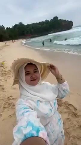 A woman in a white hijab and a large straw sunhat poses for a selfie video on a beach. She is standing on the sand with gentle waves lapping nearby. In the background, other people are wading in the water. To the left, there is a headland covered in trees. The sky is overcast. A man walks behind the woman on the left side of the frame.