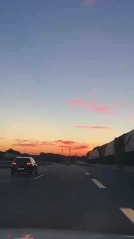 The video captures a view from a moving vehicle on a highway at dusk. The sky transitions from a deep blue at the top to shades of orange, yellow, and pink near the horizon, indicating the setting sun. Wispy pink clouds are visible. Several cars are driving on the highway in the same direction as the camera vehicle. The highway has multiple lanes marked with white lines. On the right side of the highway, there is a concrete barrier with some greenery growing on top. On the left side, there are trees and some distant structures, possibly including wind turbines. The overall mood is serene and atmospheric, characteristic of a sunset drive.