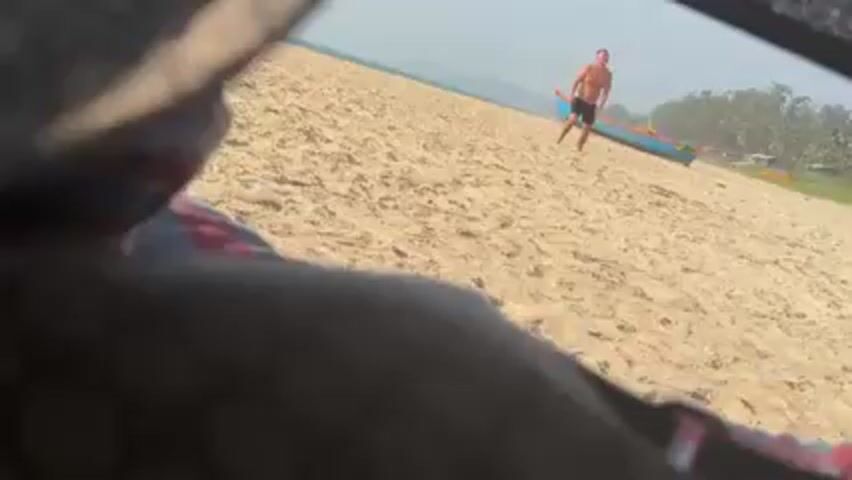 From a first-person perspective inside a vehicle, a shirtless man in dark shorts runs across a sandy beach toward a light blue boat. In the background are green trees and other colorful boats. A woman in a white swimsuit is walking in the background.