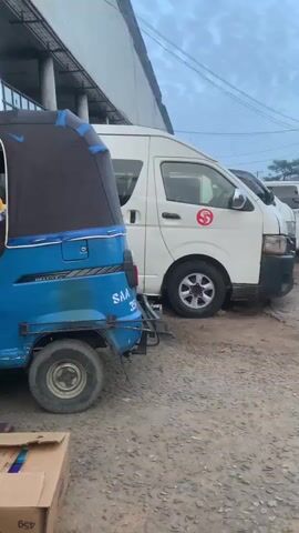 A man wearing a yellow shirt and black pants walks past a blue auto-rickshaw and two white vans. He is carrying a circular tray laden with items on his head. Another man in a dark blue shirt and black pants appears to be placing items into the side of one of the white vans. The ground is dirt and gravel. The sky is overcast.