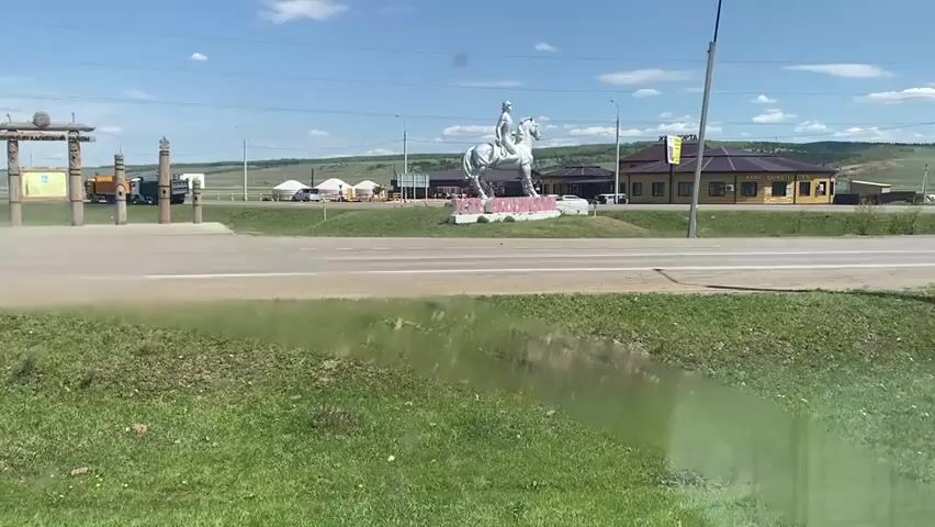 The video shows a rural intersection with a road running horizontally across the frame. In the center of the intersection is a traffic circle with a statue of a man on a horse and the name "ZABAIKALYE" in pink letters. To the left of the traffic circle is a decorative gate made of wood and stone. Behind the gate, there is some construction equipment. To the right of the traffic circle is a tan building with a sign that reads "KAPE CAHHOE CTEKNO". In the background, there are green hills and a clear blue sky with some clouds. A blue Toyota C-HR drives along the road, kicking up dust as it passes. There are also power lines and street lights visible in the background. The foreground is filled with green grass.