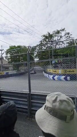 This is a vertical shot from behind a fence of a go-kart race. Several people are visible in the foreground, their backs to the camera, wearing hats and casual clothing. Beyond them, a go-kart track is visible with safety barriers and advertising. A go-kart is seen rounding a corner on the track, followed by another. Trees and a cloudy sky are visible in the background.
