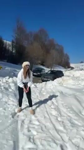 A woman is outdoors in the snow on a bright, sunny day. She is wearing a white sweater, black leggings, and a white knit hat. There are two cars parked nearby, and a telephone pole is to her right. She attempts a high kick, then stumbles and falls.