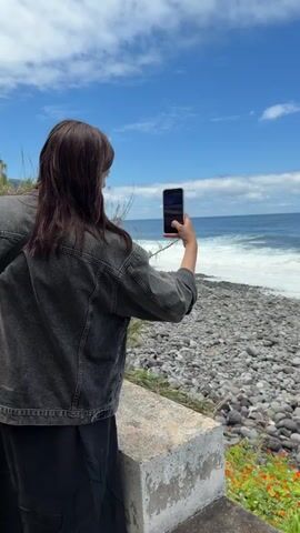 A person is taking a picture of a pebble beach and ocean with a smartphone. The person is standing on a wall next to some vegetation. They are wearing a gray denim jacket and black skirt. The pebble beach slopes down to the water, and the ocean is a dark blue with white-capped waves. The sky is blue with scattered clouds.
