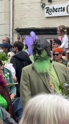 The video captures a vibrant scene in front of a building labeled "Roberts Curiosities". A crowd of people are gathered, several holding up phones, suggesting they are recording or taking pictures. Focus is drawn to a man in the foreground with his face and beard painted a bright green. He wears a pilot's helmet and goggles, a green shirt, floral tie, and a tweed jacket. To his right, a young girl with a flower crown is perched on someone's shoulders, looking towards the crowd. The overall atmosphere is festive and celebratory.