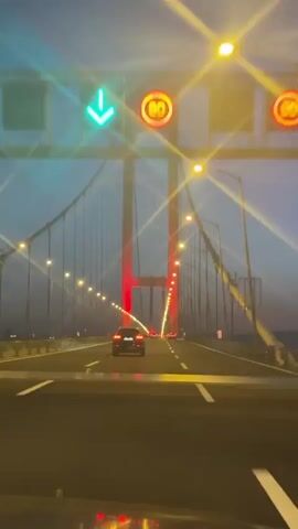 The video shows a car driving across a suspension bridge at dusk. The bridge is lit by streetlights along the roadway, and the support pillars are illuminated in red. Other cars can be seen on the bridge. The sky is a dusky blue. The viewpoint is from inside the vehicle, looking forward.