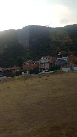 The video shows a side view of a moving vehicle. In the foreground, there is dry, yellow grass and a sidewalk. In the middle ground, there are several houses with red tile roofs and solar panels, as well as some trees and bushes. In the background, there is a large, green, forested mountain with a wind turbine at the top. The sky is overcast. A blue road sign is visible on the right side of the frame, with white text and an arrow pointing to the left.
