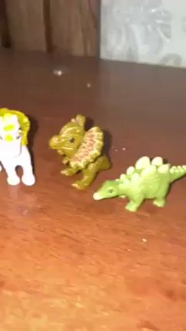 The video shows three small toy figurines on a wooden surface. The figurine on the left is a white horse with a yellow mane and tail. The other two figurines are green dinosaurs. The first dinosaur is a Dilophosaurus, standing with its frill visible. The second dinosaur is a Stegosaurus. The wooden surface appears to be a table or desk. A wall is visible in the background.