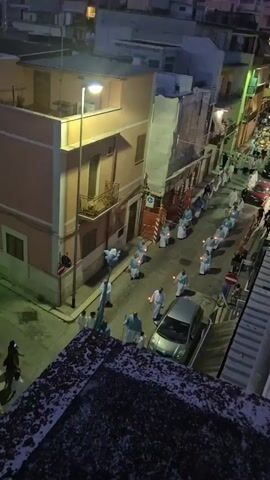 The video shows an aerial view of a street at dusk. A procession of people, many wearing white robes and holding candles, walk down the center of the street. They are organized in a line. There are buildings on either side of the street, some with balconies. Cars are parked along the street and some are in the procession. A few people are standing on the side of the street observing the procession. The sky is a deep blue, indicating that it is evening. The lighting appears to be a mix of streetlights and the light from the candles. The overall mood of the scene seems solemn and possibly religious.