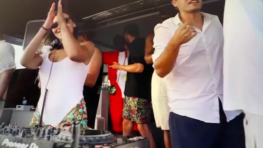 The video shows a DJ set on what appears to be a boat. In the foreground, a female DJ in a white swimsuit with a colorful sarong is operating a Pioneer DJ controller, wearing headphones and sunglasses. She is smiling and appears to be enjoying herself. To her right, a man in a white shirt and dark shorts is dancing, also smiling and looking towards the DJ. Behind them, several other people are visible, some dancing and some talking. The atmosphere seems festive and lively. The background is somewhat blurred, but suggests an outdoor setting with bright lighting, likely during the day.