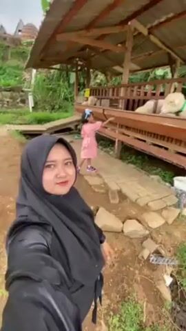 The video shows a woman wearing a black hijab standing in front of a goat pen. To her right is a young girl in a pink dress. The girl walks towards the pen, which is under a wooden structure with a roof. There are three white goats inside the pen, which is raised off the ground. A sign reads "Dilarang Memberi Makan Domba," which translates to "Do not feed the sheep." The ground is a mix of dirt, grass, and brick pathways. The area appears to be a rural setting with greenery visible in the background. The woman waves at the camera.
