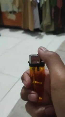 The video shows a close-up of a person's hand holding a disposable lighter. The lighter is orange and transparent, with a black cap and a metal spark wheel. The person is pressing down on the spark wheel with their thumb, and a flame ignites. The flame is bright and orange, with a blue base. The background is blurred, but it appears to be an indoor setting with tiled flooring and some hanging fabric. The video is short, lasting only a few seconds, and focuses on the action of lighting the lighter.