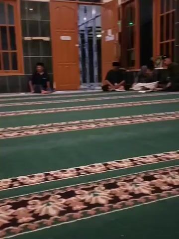 The video shows a group of men sitting on a green carpet with a patterned border inside a room. The room appears to be a mosque or prayer hall, with wooden doors and windows visible in the background. Several men are seated on the carpet, facing in the same direction. Some appear to be reading or engaged in prayer. The lighting suggests it is evening or indoors.