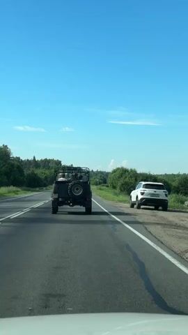 The video shows the rear view of a dark green open-top military-style vehicle driving down a paved road on a clear, sunny day. The vehicle has a spare tire mounted on the back. There are two people visible in the vehicle, one in the passenger seat and one behind the driver. A white car is parked on the right side of the road, partially obscured by bushes. The road is bordered by lush green trees and vegetation. The sky is bright blue with a few scattered white clouds. The camera appears to be from another vehicle following behind.