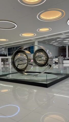 The video shows a polished metal sculpture inside a large building, likely a shopping mall or airport. The sculpture features two large, circular, dish-shaped components connected by a curved metal band. The reflective surfaces of the sculpture mirror the surrounding environment, including the tiled floor, ceiling lights, and other parts of the building. The sculpture rests on a circular base surrounded by a square glass enclosure with a black frame. The background reveals other retail spaces, ongoing construction, and people walking in the distance. The ceiling is adorned with multiple circular light fixtures.
