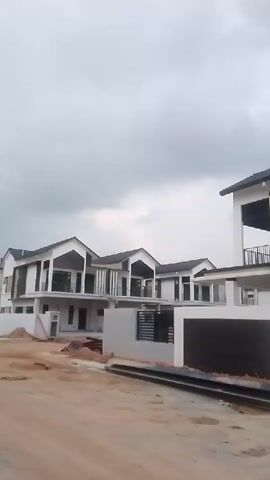 The video shows a street view of several modern houses under construction or recently completed. The houses are multi-story with white and dark brown exteriors, large windows, and dark tiled roofs. Some houses have balconies. The street in front of the houses is unpaved with dirt and construction debris. The sky is overcast. The camera is panning from left to right, showing more of the houses and the street.