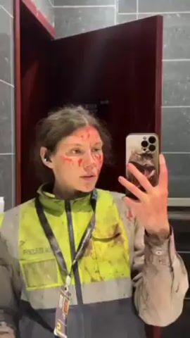 In a medium shot, a person stands in front of a bathroom mirror, taking a selfie with a smartphone. The person is covered in what appears to be blood on their face, hands, and clothing. They are wearing a high-visibility work jacket, and an identification card hangs around their neck. The person is holding a large wrench in their left hand. The bathroom has gray tiled walls and a dark red door is partially visible in the background. The person's expression is neutral, with a slight downward gaze towards the camera.