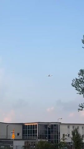 The video shows a clear sky with a few clouds and a commercial airplane flying at a high altitude. The plane is white with a visible tail. Below the plane, a building with a modern glass-fronted section and a sign that reads "METALOWIEC" is visible. A tree with green leaves is partially in the frame on the left side. A yellow traffic sign is also present near the building. The overall lighting suggests it is daytime, possibly late afternoon or early evening due to the warm tone of the light. There are no people visible in the frame. The camera appears to be handheld, with slight movement noticeable.