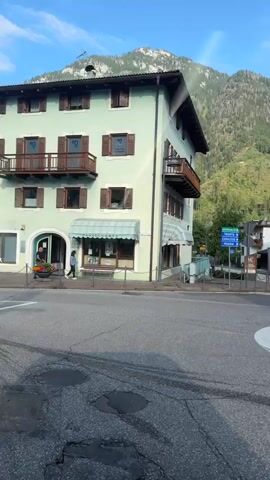 The video captures a street scene in a European town with buildings and mountains in the background. In the foreground, there's a roundabout with traffic signs. A person is seen walking on the sidewalk on the left side of the street, and another person is visible near a building on the right. The sun is shining, and the sky is blue. The buildings are multi-story structures with windows and balconies. The overall atmosphere appears peaceful and serene, with a hint of activity from the pedestrian.