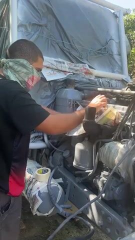 A person wearing a mask and a head covering is working on the engine of a truck. The engine and surrounding areas are covered in gray primer and protective plastic sheeting. Tools like masking tape and what appear to be brushes or applicators are visible. The environment appears to be outdoors, with trees and sunlight visible in the background.