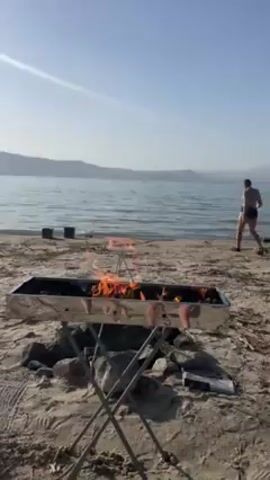 The video shows a sandy beach with a fire pit in the foreground and a woman wading into the water in the background. The fire pit is constructed of metal and is raised off the ground on a frame. Orange flames are visible within the pit. The woman is wearing dark swimwear and walks into the water. The water is calm with gentle ripples. In the distance, there are hills or mountains under a light blue sky. There are some dark objects placed on the beach at the water's edge. The light is bright and sunny. The camera is stationary.