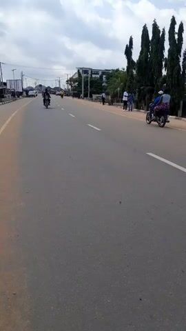 The video shows a street scene with a paved road and a dirt shoulder. On the road, there are several motorcycles and cars in motion, and people walking on the sidewalk. Buildings line the street on the left side of the frame. The sky is cloudy.