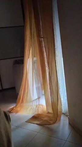 A sheer, golden curtain hangs in front of a window, allowing bright light to filter through. The curtain pools on the tiled floor. To the left of the curtain, a tall, white appliance with vents is visible. The room appears to be dimly lit, with shadows cast by the curtain and the appliance.