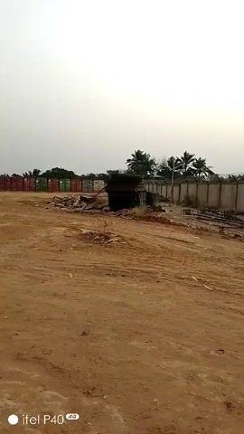 The video shows a dirt lot enclosed by concrete walls topped with barbed wire. There's a metal gate with a concrete post. Palm trees are visible in the background. Several large vehicles are parked in the distance beyond the gate. Debris and discarded items are scattered around the lot. One person is briefly visible walking near a small shed. The sky is overcast.  The footage appears to be taken with a mobile phone.