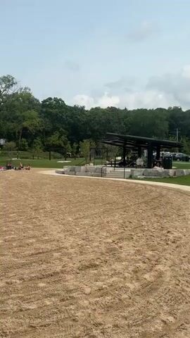The video opens on a wide shot of a sandy beach with tire tracks visible in the sand, suggesting recent activity. In the distance, there is a treeline, a grassy area, and a modern-looking structure with a dark roof, possibly a pavilion or cafe. Several people are scattered across the grassy area, some sitting under umbrellas and others lying on blankets. A few more people are seen closer to the water's edge. The scene then pans to the left, revealing a body of water with a few individuals in it. The camera continues to pan left, showing more of the beach and the water. As the video progresses, the perspective shifts to a view closer to the water, with a rocky embankment separating the sand from the lake. Several people are seen on paddleboards in the water. The video concludes with a view of the lake and the surrounding shoreline. The sky is mostly clear with a few scattered clouds. The overall impression is a relaxed, recreational setting on a sunny day.