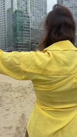 A person in a yellow shirt and brown shorts is on a sandy beach, performing a handstand. The person is facing away from the camera and their legs are in the air. Other people are visible in the background, some walking and some sitting on the sand. Tall buildings are in the background, suggesting an urban environment.