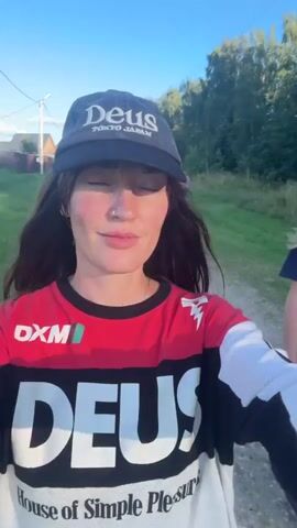 The video shows a woman in the foreground, wearing a black Deus Ex Machina baseball cap and a long-sleeved jersey with red, white, and black panels, also featuring the Deus logo. She is smiling and looking at the camera. Her long dark hair is visible. Behind her, a path leads into a grassy area with trees. In the background, two other individuals are walking away from the camera. The person closer to the woman is wearing a dark t-shirt. The person further back is wearing a grey sweatshirt and bright yellow pants. The environment appears to be rural, with a blue sky and some vegetation visible. The woman speaks in Russian, mentioning a large empty field near their dachas two years ago.