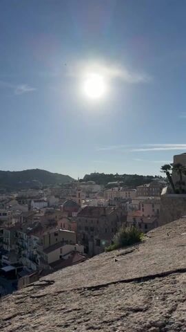 The video presents a slow, panoramic view of a picturesque coastal town and its harbor on a bright, sunny day. The scene opens with a wide shot of the town, characterized by buildings with terracotta roofs, nestled against a backdrop of rolling hills and mountains. The sun is high in the sky, appearing slightly to the right of the center, casting a strong glare and illuminating the scene. A prominent building with a red-tiled dome is visible among the town's structures.

As the camera slowly pans to the right, more of the town's dense architecture is revealed. The pan continues, gradually bringing a large, active harbor into view. The deep blue waters of the harbor are filled with numerous boats, including yachts, docked at several long piers. The town stretches along the harbor's edge, blending into the landscape. The rugged mountains continue to form a dramatic backdrop, extending along the coastline. The sky is clear and blue with a few scattered white clouds. The foreground throughout the video consists of a textured, rough stone surface, indicating the recording was made from a high vantage point, possibly a defensive wall or a natural rock formation overlooking the entire bay. The video ends with a comprehensive view of the harbor, town, and mountains under the bright daylight.