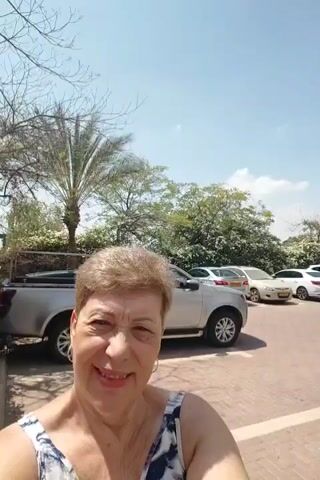 A woman is taking a selfie outdoors. She is smiling and looking at the camera. Behind her, there is a flowering bush with pink and red flowers, a silver truck, and a tall building. The sky is blue and clear.