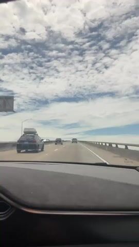 This is a first-person view from inside a car driving on a highway. The car has a dashboard in the foreground. The road is elevated and appears to be a bridge, with barriers on the right side. Several other cars are visible on the road ahead, some in the same lane and some in adjacent lanes. The sky is filled with dramatic, scattered white clouds against a blue sky. The sun appears to be shining, casting light on the scene. The overall impression is a journey through an open landscape under a dynamic sky.