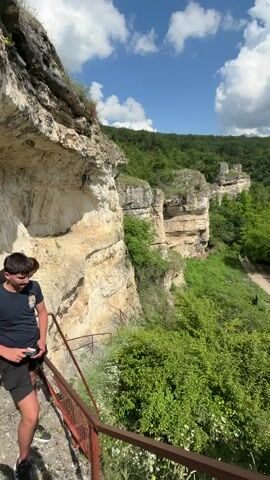 A person standing on a metal railing-enclosed viewing platform overlooks a valley with steep, rocky cliffs and abundant green foliage. A dirt road winds through the valley, where a black car is visible. The sky is bright blue with scattered white clouds.