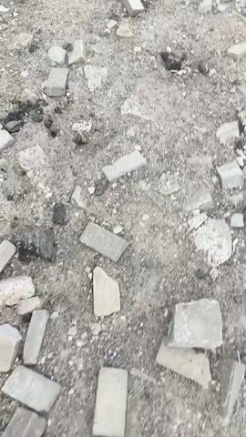 This is a short, shaky video with a neutral color palette, filmed from a first-person perspective, likely outdoors in daylight. The scene is a ground covered with scattered debris, primarily broken concrete or stone pieces, along with some dirt and small pebbles. Some larger, flat rectangular pieces, possibly remnants of paving stones or bricks, are also visible. The texture of the ground suggests an unkempt or construction-related environment.

In the bottom left corner of the frame, a person's legs and feet are visible, wearing grey sneakers with white laces and a blue accent on the heel. The person appears to be walking through the debris. The movement of the camera suggests the person is navigating the uneven terrain.

There are no discernible people other than the one whose feet are visible, no animals, and no explicit objects that are clearly identifiable beyond the general debris. The setting appears to be a damaged or unfinished outdoor area, possibly a demolition site, a neglected pathway, or an area undergoing renovation.

The activity is simply walking. The time of day appears to be daylight, given the lighting, and there are no clear indicators of weather conditions. The location context provided is Den'kovo, Russia, which does not offer specific visual cues within the video itself. No text is visible in the frame. The overall mood is one of mundane movement through a somewhat disheveled environment.