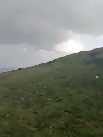 The video captures a panoramic view of a lush green mountainous landscape under a cloudy sky. The camera pans across rolling hills covered in short, vibrant green grass, interspersed with occasional wildflowers. In the distance, layers of hazy, darker green mountains recede into the horizon, suggesting a vast and expansive terrain. The sky is overcast with thick, grey clouds, creating a diffused light that softens the overall scene. There are no clear signs of human activity or specific objects in the foreground or middle ground. The video focuses on the natural beauty and ruggedness of the mountainous environment, with a sense of solitude and openness.