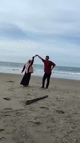 The video shows a couple on a sandy beach with the ocean in the background. The sky is cloudy. A piece of driftwood is lying on the sand in the foreground. A man in a red shirt and black pants is standing on the right, facing the camera. A woman in a pink hijab and a dark dress is on the left, also facing the camera. They are holding hands and appear to be dancing or posing for a photo. The man has his left hand on his hip. The woman is leaning back slightly. The man then turns and walks away from the camera towards the right side of the frame. The woman remains in place for a moment before the video cuts off.