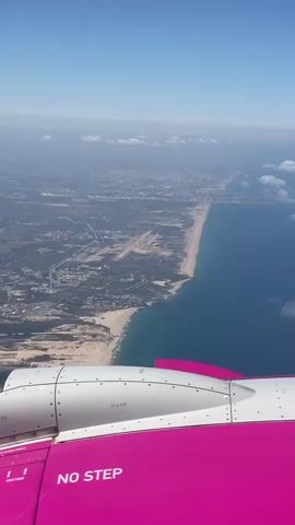 The video shows an aerial view from the window of an airplane. In the foreground, a portion of the airplane's wing and engine nacelle are visible. The wing is painted bright pink and has the text "NO STEP" printed on it. The engine nacelle is predominantly white with some metallic detailing.

The background reveals a coastline with a sandy beach transitioning into the turquoise ocean. A city or urban area stretches inland from the coast, with a noticeable runway or airfield visible near the coastline. The sky is clear and blue, with some scattered clouds appearing hazy due to the altitude and atmospheric conditions. The overall impression is that of an airplane in flight, approaching or flying over a coastal city.
