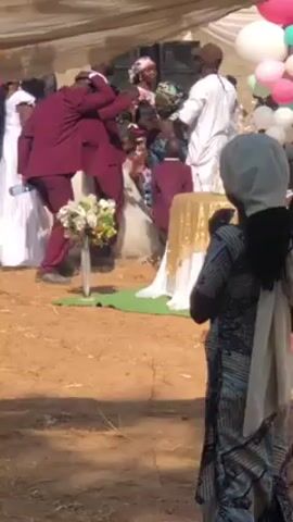 The video depicts an outdoor wedding scene. A bride in a white wedding gown is accompanied by two men in maroon suits. A small child, also in a maroon suit, stands near a table draped in gold fabric. Several people, including guests and staff, are seen around the area. Some individuals are wearing traditional attire, while others are in casual clothing. A white tent-like structure provides shade over the wedding area, and balloons in pink, white, and green decorate the scene. The ground appears to be dry and dusty, and the overall atmosphere seems festive and celebratory.