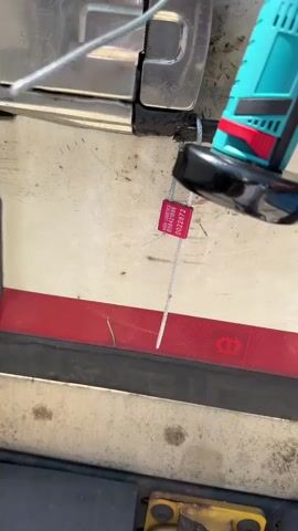 A pink tamper-evident seal with the number 0022872 is attached to a metal cable, which is connected to a trailer door. A teal and black tool is positioned near the cable. The trailer door is partially open. The surface of the trailer is dirty and stained.