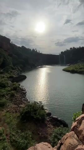 A vertical video shows a wide shot of a reservoir with two waterfalls cascading down in the distance. The sun is high in the sky, casting a shimmering reflection on the water. The foreground is a rocky, overgrown embankment with sparse green bushes. The overall atmosphere is serene and natural.