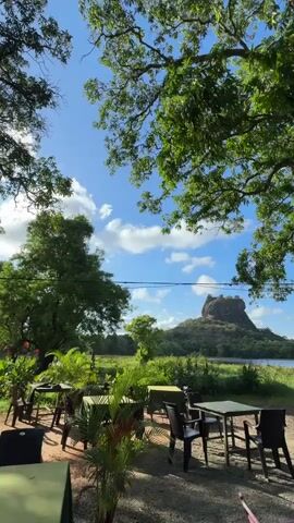 The video shows a scenic view of Sigiriya rock fortress in Sri Lanka from what appears to be an outdoor seating area. The fortress stands prominently in the distance, surrounded by lush greenery and a body of water. In the foreground, there are tables and chairs under the shade of large trees, creating a relaxed, outdoor dining setting. A swing hangs from one of the tree branches, adding to the peaceful atmosphere. The sky is blue with scattered white clouds, suggesting a clear, sunny day. No people are visible in the scene. The overall impression is that of a tranquil and picturesque location, perfect for enjoying the natural beauty of the landscape.