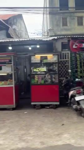 The video captures a street scene with several businesses. In the first segment, a small shop labeled "BEREUKAT" is visible, stocked with various goods. A motorcycle is parked outside. The camera then pans to a narrow alleyway lined with houses and vegetation. Another segment shows a blurry view of a street with more shops and people. One shop appears to be a repair service with several motorcycles inside and people working on them. The overall impression is of a busy, urban environment with small businesses and residential areas.