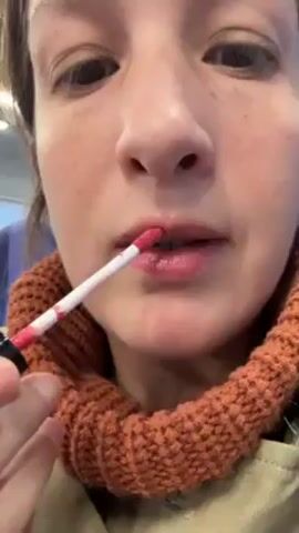 A woman is applying pink lip gloss in an indoor setting. She has brown eyes, fair skin with freckles, and is wearing an orange knit turtleneck sweater. She holds the lip gloss applicator with her right hand, focusing on her upper lip. The background is slightly blurred, suggesting an interior space, possibly a vehicle or waiting area.