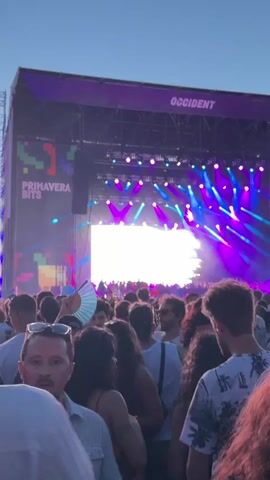 A low-angle medium shot captures a large crowd of people standing close together at an outdoor music festival during the daytime. The camera pans from left to right, showing the faces of various individuals in the foreground, and the backs of others in the middle ground, who are mostly focused on the stage in the distance. The stage is visible at the top of the frame, with large video screens displaying "PRIMAVERA SOUND" and "OCCIDENT" in white text on a dark background. Bright purple and blue lights illuminate the stage, and large speakers are mounted on either side. The sky above is clear and blue.