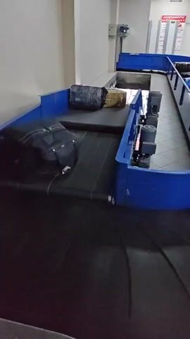 The video shows a baggage carousel in an airport or transportation hub. The carousel is primarily blue with a black conveyor belt. Several pieces of luggage, including black suitcases and wrapped packages, are moving along the belt. The carousel snakes around, with a view of the inner workings of the belt system, including the motors, in one segment.  The environment looks clean and brightly lit, with a tiled floor, white walls, and signage visible in the background. The scene conveys an empty or quiet state of the baggage claim area.
