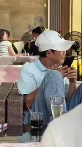 Here is a detailed description of the video:

The video shows a man sitting in a wicker chair at what seems to be an outdoor cafe. He wears a white baseball cap with "LA" embroidered on it, a light blue polo shirt, and blue jeans. He is looking down at a smartphone, engrossed in something on the screen. His legs are crossed. Beside him on a small mirror-topped table are two glasses - one with a dark liquid and a straw, and another with a light-colored liquid, possibly a beverage.

In the background, other people are sitting at tables. A couple is seen in conversation, and another person is partially visible behind them.  A small electric fan is also visible near the man, suggesting warm weather. The cafe has a relaxed ambiance with open-air seating and textured concrete walls.