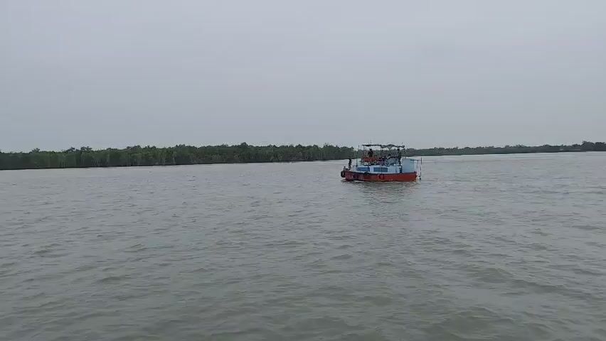 A wide shot captures a boat navigating a body of water. The boat is red and white with a blue roof. Several people are visible on the deck of the boat. The background features a line of trees, possibly a mangrove forest. The sky is overcast, and the water appears calm with slight ripples. The boat is moving from left to right in the frame.
