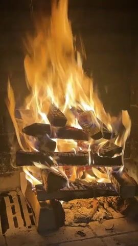 A roaring fire is burning in a fireplace. The fire is large and the flames are orange and yellow. The wood in the fire is stacked in a criss-cross pattern. The fireplace is made of brick. There is ash and embers beneath the burning wood.